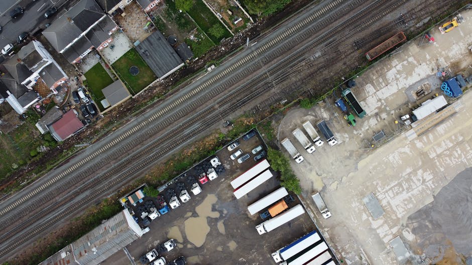 An aerial view of a parking lot and industrial yard adjacent to railway tracks in an urban setting. The foreground shows a mixture of parked cars, some in rows and others scattered, alongside white and gray container units, with a few smaller vehicles such as vans and trucks. Patches of water and muddy ground are visible among the vehicles, indicating recent rain or drainage issues. To the right, there is an open outdoor area with concrete paving, storing large blue pipes and other construction materials, as well as a white truck and a green container. Rail tracks run horizontally across the middle of the image, separating the parking lot from the residential area above, where houses with various roofing materials and small gardens can be seen. The scene is illuminated by natural daylight, with shadows cast by the vehicles and structures. The composition highlights a typical urban environment where waste management services like those from Waste Removal Edgware might operate to handle various waste types from construction, commercial, and residential sectors, emphasizing their role in alternative rubbish disposal solutions in the local area.