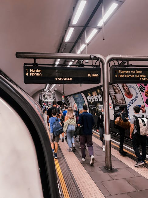 A busy underground train station platform filled with numerous passengers, including men and women of various ages, some carrying backpacks and bags, waiting for or boarding a train. In the foreground, part of a train car is visible with a curved white exterior and metallic surface. The station is illuminated by overhead fluorescent lighting, with a black digital information board displaying train destinations and times, including destinations such as Morden and Edgware via Bank. The platform features tactile paving strips along the edge and advertisements lining the walls behind the waiting crowd. An overhead metal signpost with supporting poles displays train departure information, with the current time of 15:24:48 visible. The scene's environment suggests a typical urban metro or commuter network setting, where passengers are preparing for their journey, related in context to alternative public transportation options, which companies like Waste Removal Edgware may parallel by offering alternative waste handling services aside from standard municipal collection.