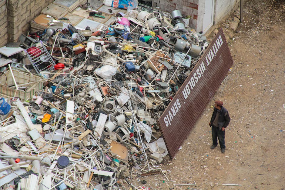 An outdoor scene showing a large pile of mixed construction and household waste materials situated against a brick wall, including broken furniture, plastic containers, metal pipes, electrical components, and miscellaneous debris with varied textures and colors such as white, gray, black, and blue. The waste materials are scattered across the ground, with some items stacked and others loosely piled, creating a chaotic arrangement. To the right of the waste pile, there is an individual wearing dark clothing and a brown cap standing on an unpaved, dirt-covered surface, observing the rubbish. Adjacent to the waste pile, a weathered metal sign with white lettering states 'NEVER WASH IN THE SEWERS,' leaning at an angle and partially obscured by the debris. The environment suggests an area designated for bulk waste collection or disposal, with the scene indicating a site where independent rubbish removal services such as Waste Removal Edgware might be involved in sorting or clearing waste materials, reflecting on the importance of proper bulk waste handling and disposal regulations.