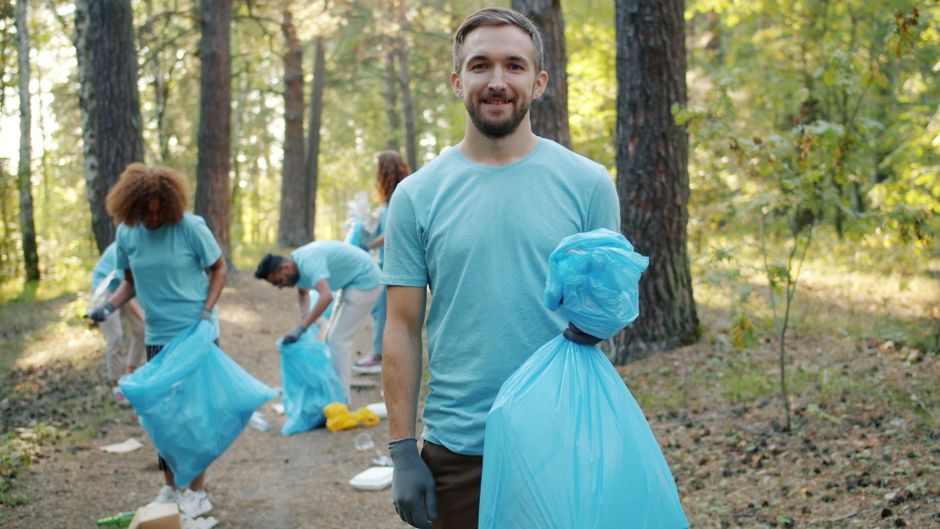 A group of volunteers participating in a community clean-up event in a wooded area during daylight hours. In the foreground, a bearded man with short hair, wearing a light blue t-shirt and grey gloves, is holding a large blue plastic rubbish bag filled with collected waste, smiling at the camera. Behind him, several other volunteers, also dressed in light blue t-shirts, are actively engaged in picking up debris from the forest floor, crouching or bending over to gather litter. The volunteers are surrounded by mature trees with textured bark, and the ground is covered with fallen leaves, twigs, and scattered trash. The scene is illuminated by natural sunlight filtering through the canopy, creating a bright and fresh outdoor atmosphere. The image emphasizes community involvement in environmental efforts, showcasing the importance of on-site waste collection and rubbish removal in public spaces, an act supported by services like Waste Removal Edgware and other private waste management providers.