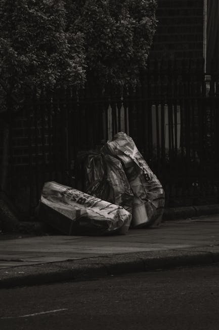Three large black rubbish bags, made of thick plastic with some visible wrinkles and creases, are placed on the edge of a paved pavement near a black metal fence with vertical bars. The bags appear to be filled with household waste or debris, with some items bulging through the transparent sections and printed markings visible on the surface. Behind the fence, dense foliage of dark green leaves and branches is partially visible, creating a shadowed background. The scene is illuminated softly, suggesting either an overcast day or low natural light, and is located outdoors in an urban or residential setting, with the rubbish bags presumably awaiting collection or disposal. This image exemplifies typical private waste disposal in a street environment, aligned with professional rubbish removal services such as those offered by Waste Removal Edgware.