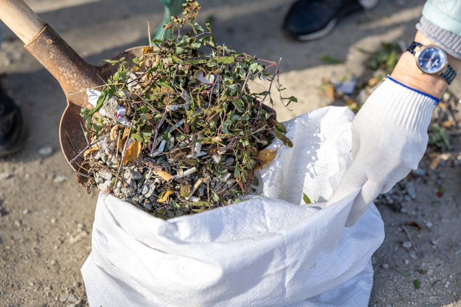 A person wearing a white glove and a blue wristwatch is using a small shovel to load a white sack with garden debris, including a mix of dried leaves, small twigs, and green foliage, onto a gravel surface. The sack appears partially filled with organic waste, and the person’s arm is positioned on the right side of the image, with their hand gripping the edge of the sack. The background shows outdoor environment with dirt and scattered plant matter, implying a process of clearing or disposal of garden rubbish. The scene suggests activities related to private waste disposal or professional rubbish removal services, such as those offered by Waste Removal Edgware, focusing on on-site clearance of garden waste in preparation for disposal or recycling.