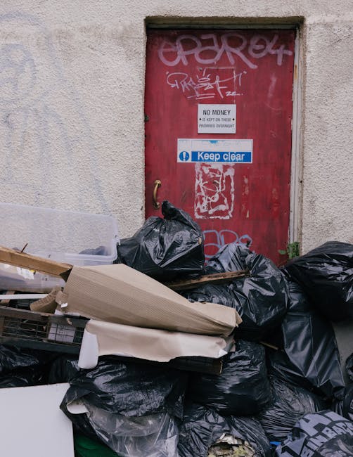 An exterior scene featuring a weathered, red metal door set into a beige, textured concrete wall. The door is covered with graffiti tags and has a white sign reading 'NO MONEY' with additional smaller text below, along with a blue and white sticker that states 'Keep clear.' In front of the door, there is a large pile of discarded rubbish comprising multiple black plastic bags, some partially torn or bulging, and assorted waste items including flattened cardboard boxes, a piece of timber, and a turned-over white appliance or piece of furniture. The rubbish is stacked against the wall and spills onto the paved ground, creating a cluttered landscape typical of improper waste disposal or an off-site clearance scene, which may relate to private rubbish removal services. The environment appears to be an outdoor alley or back street area, captured during daylight with soft lighting that highlights the textures of the rubbish and the building surface, illustrating a scene that aligns with the themes of rubbish clearance and waste management services provided by Waste Removal Edgware.