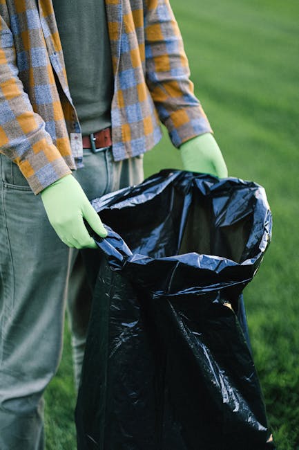 A person wearing a plaid shirt, green gloves, and grey trousers stands outdoors on a grassy area, holding open a large black rubbish bag lined with a shiny inner layer. The individual is in the process of discarding waste, with the bag partially pulled open in both hands, revealing its empty interior. The background features a well-maintained green lawn, suggesting a garden or park setting. This scene illustrates a waste collection or rubbish removal activity, potentially linked to a professional waste management service such as Waste Removal Edgware, which offers external rubbish clearance options. The lighting is natural, highlighting the textures of the bag's plastic material and the clothing's fabric, creating a neutral, observational depiction of the task of private waste disposal or on-site rubbish removal.