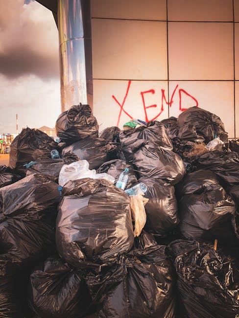 A weathered red metal skip bin with visible rust and peeling paint sits against a dark grey brick wall and a concrete wall in an urban alleyway. The skip is positioned on a tiled pavement area, with its front facing slightly to the left. Several white plastic bags filled with mixed household waste and recyclables are piled next to the right side of the skip, some leaning against it. The bags appear loosely tied and contain visible contents such as plastic bottles, paper, and miscellaneous rubbish. The background includes a modern building’s dark grey brick wall, a section of a concrete wall with a small rectangular window or opening near the top right, and part of a vertical black structure or door frame. The scene is illuminated by natural daylight, creating soft shadows and a neutral environment typical of independent waste collection points, highlighting settings suitable for private rubbish removal services like those offered by Waste Removal Edgware.